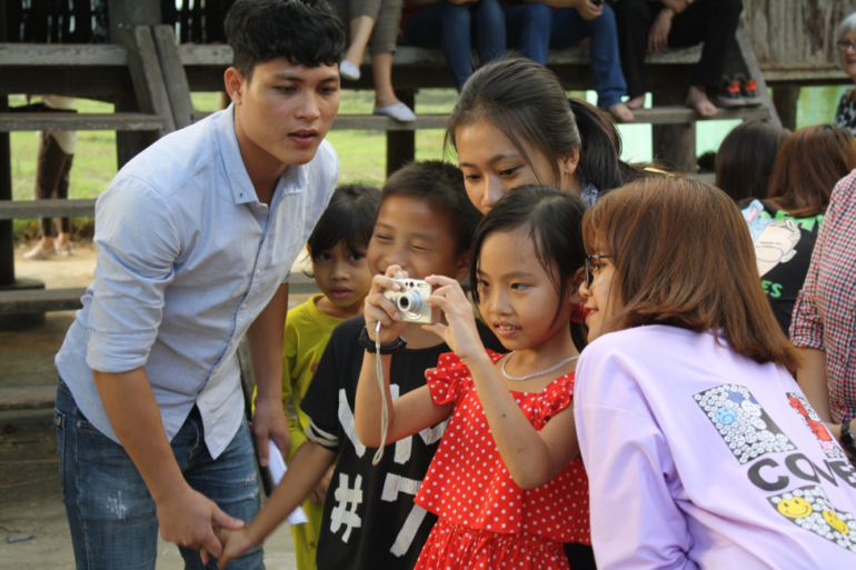 A vietnamese group of six people, both children and adults, are looking at a young girl taking a picture with a camera